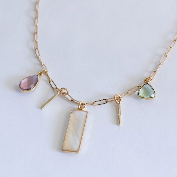 Product photo of the Athena Necklace, a gold tone paperclip style chain with two gold tone thin stick charms and three gemstone charms made of purple synthetic morganite, mother of pearl, and green chalcedony.