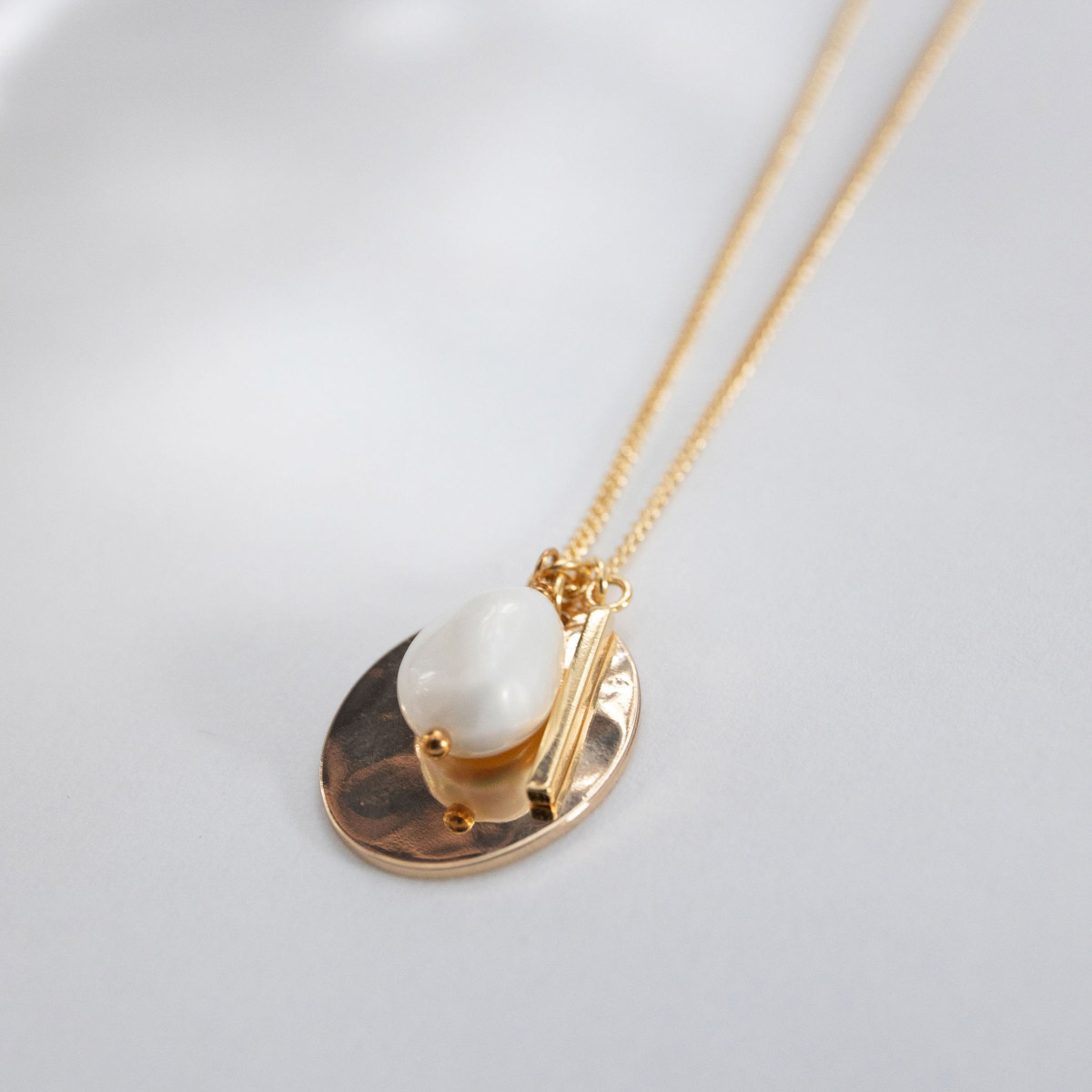 The Lady of the Empyrean Necklace, a white baroque pearl and tiny gold stick charm over a hammered oval pendant, strung on a dainty curb chain.