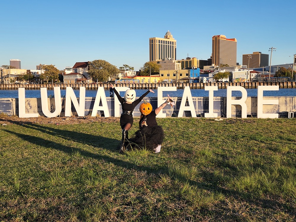 Two women dressed in black and wearing jack-o-lanterns on their heads, posing in front of a white "Lunar Faire" sign.