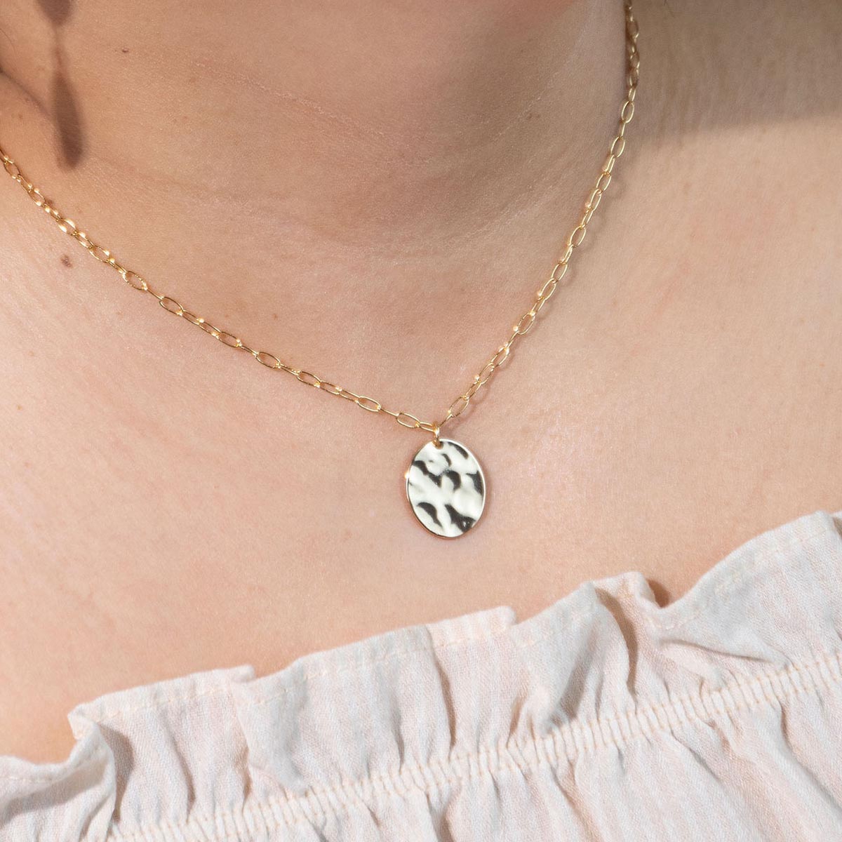 Closeup of a woman's neck modeling the Meridian Necklace, a gold hammered oval pendant on a dainty paperclip chain.