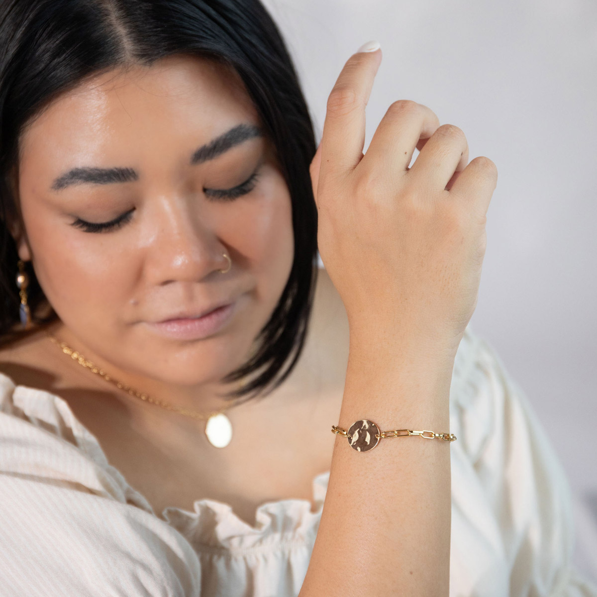Woman in a tan blouse modeling the Meridian Bracelet, a hammered gold oval charm on a thick paperclip chain.