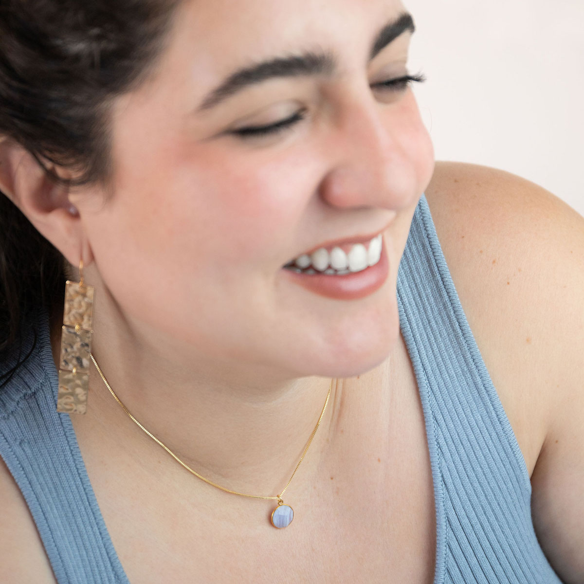 A smiling woman modeling the She Rules the Sky Necklace, a bezel-set round faceted blue lace agate on a dainty box style chain.