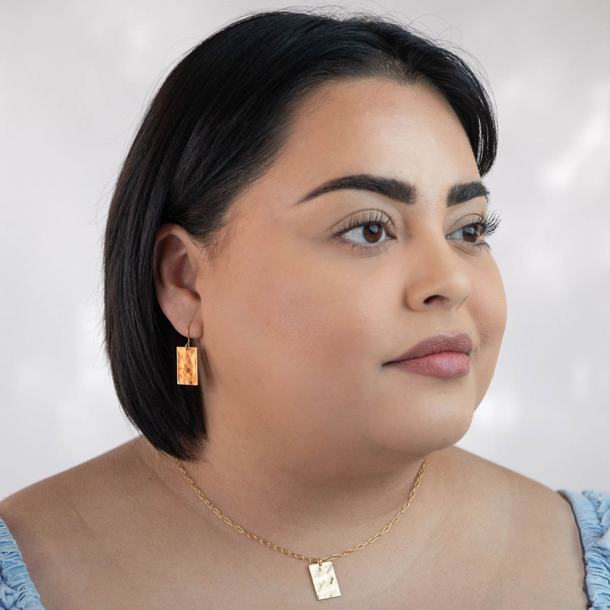 A woman in a blue blouse modeling the Zenith earrings, a gold tone hammer textured rectangle charm finished on french hooks.